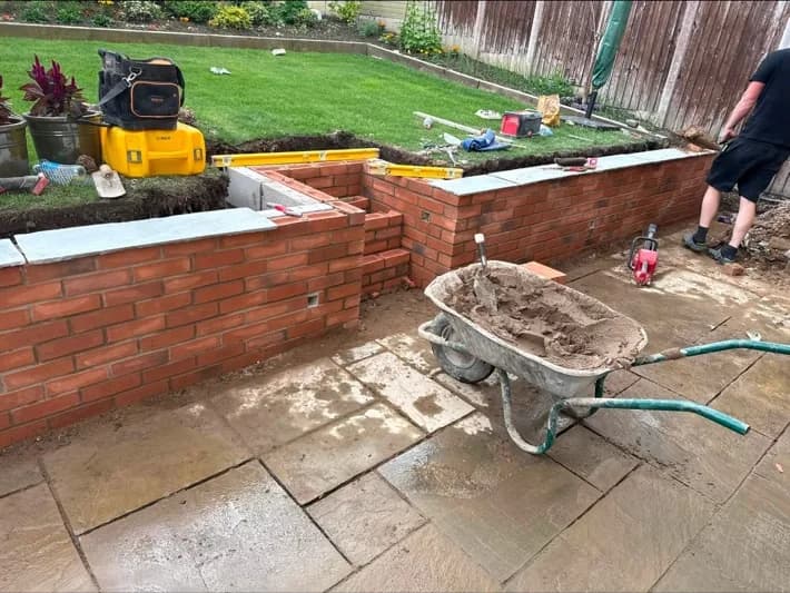 A red-brick garden wall being built with coping stones set along the top, in a domestic back garden